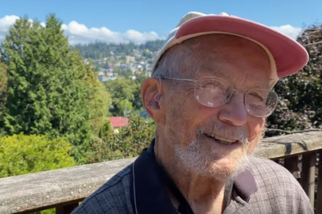 Myrl Beck smiles at the camera from his deck; behind him are the house-covered hills of Bellingham and blue sky and clouds