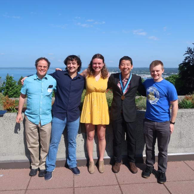 A group of five people is posing for a photo on a rooftop. The men are wearing casual clothes, while the woman is dressed more formally. The people are all smiling. In the background, there is a view of Bellingham and Bellingham Bay, looking north.