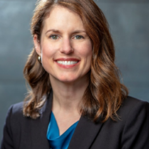 Pam Brady against a gray background, wearing a black suit over blue silk blouse