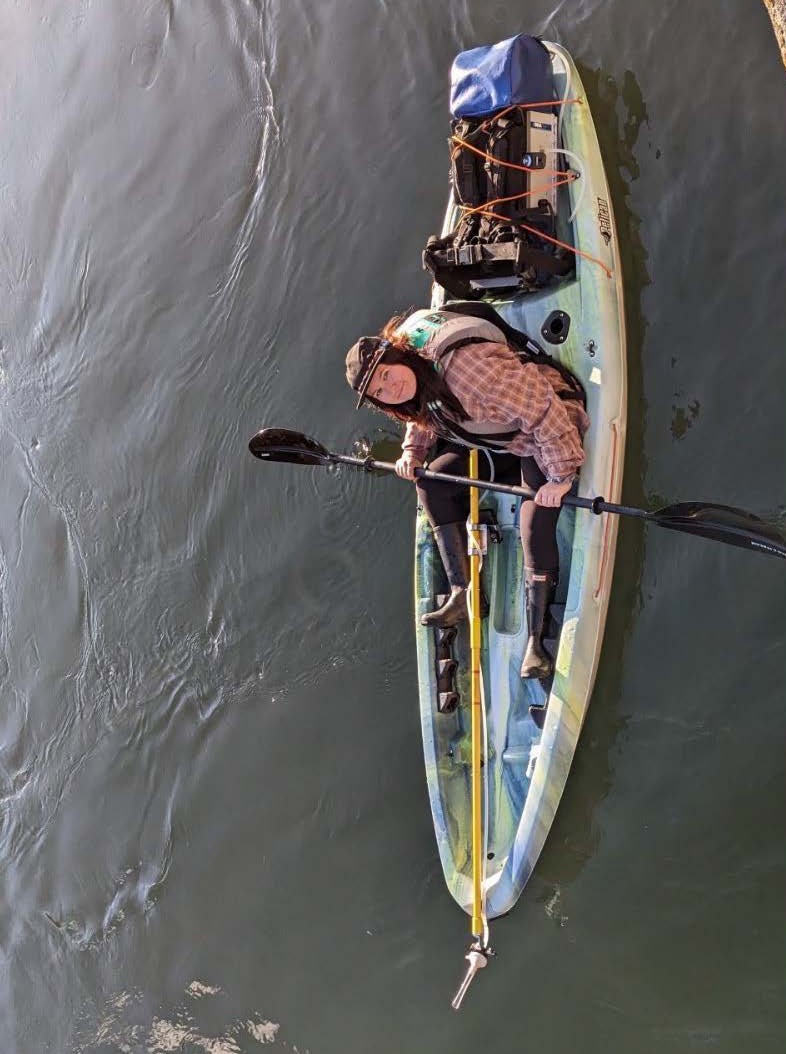 Victoria Vinecke in a kayak where the eDNA Sampler is secured at the back and tube with filter is connected to a long yellow rod.
