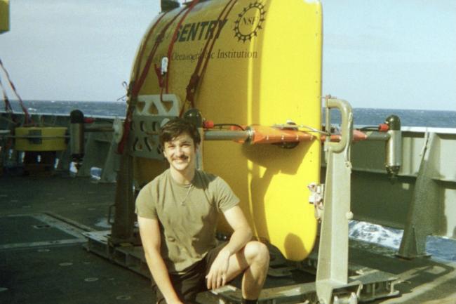 Dexter Davis on a research ship with a remotely piloted submarine behind him.